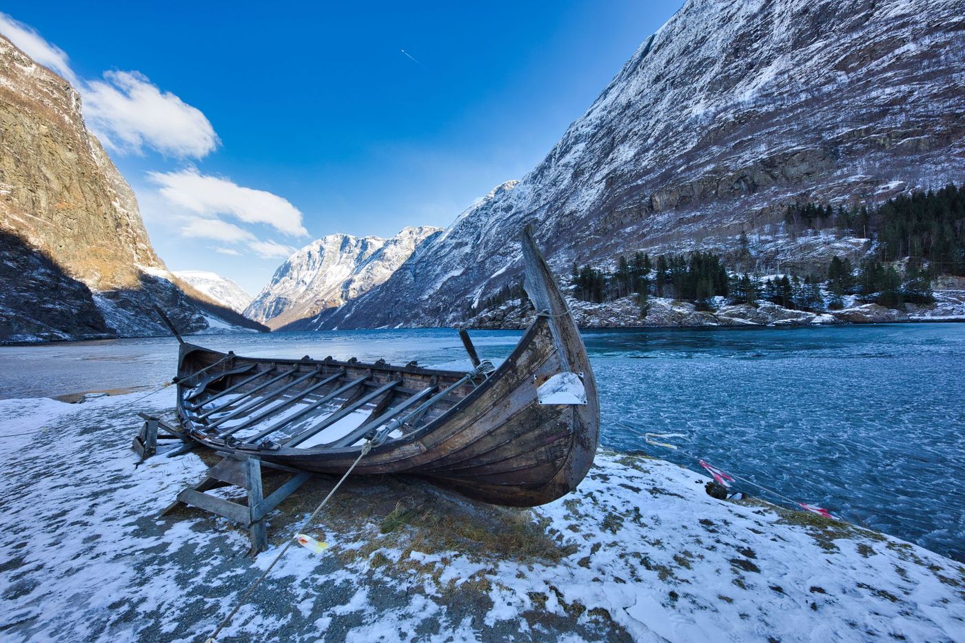 Snow-covered wooden boat on a frozen lakeshore with mountains and blue water under a bright sky
