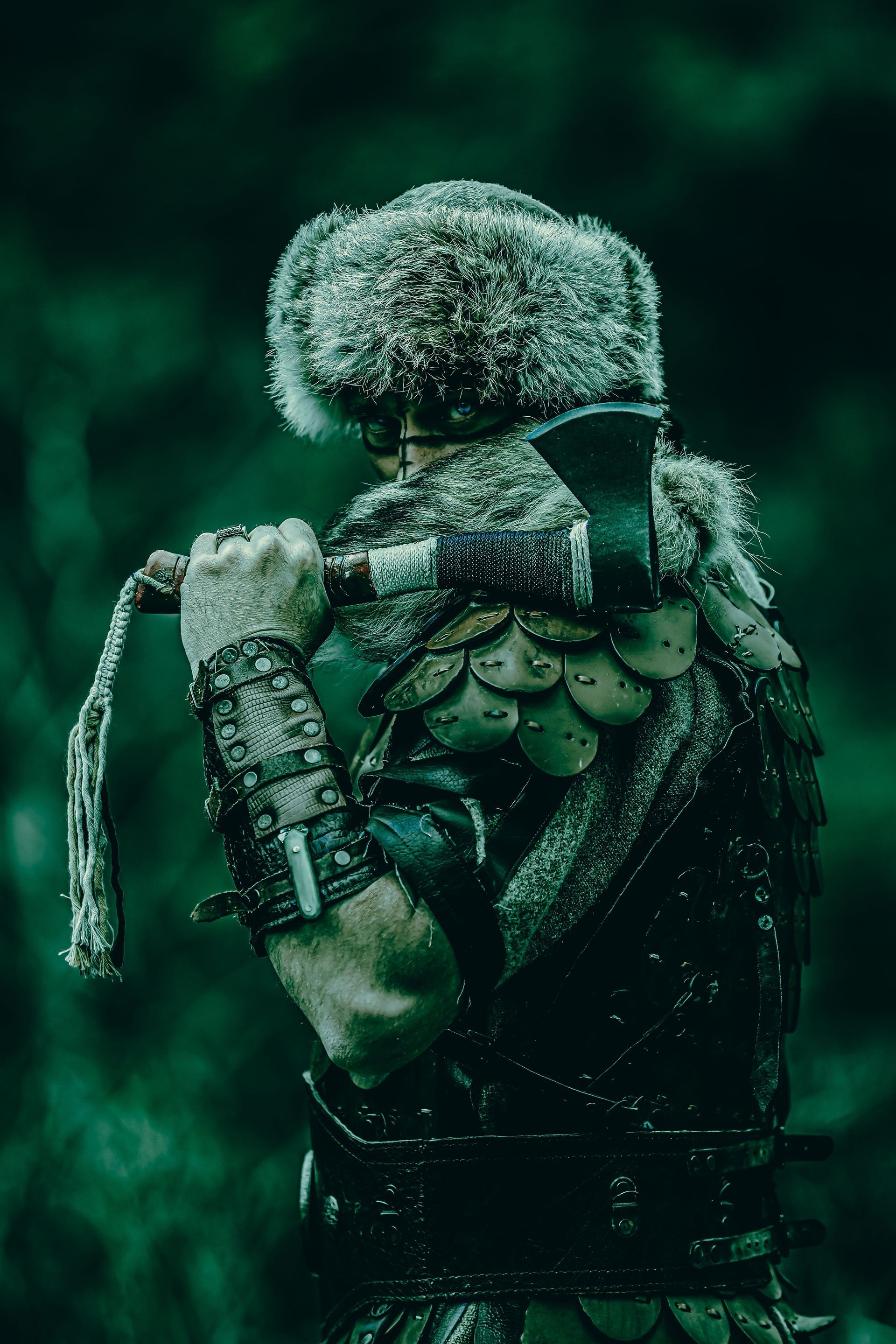 Backpacker in a fur hat and green cloak, seen from behind in a dark forest setting.
