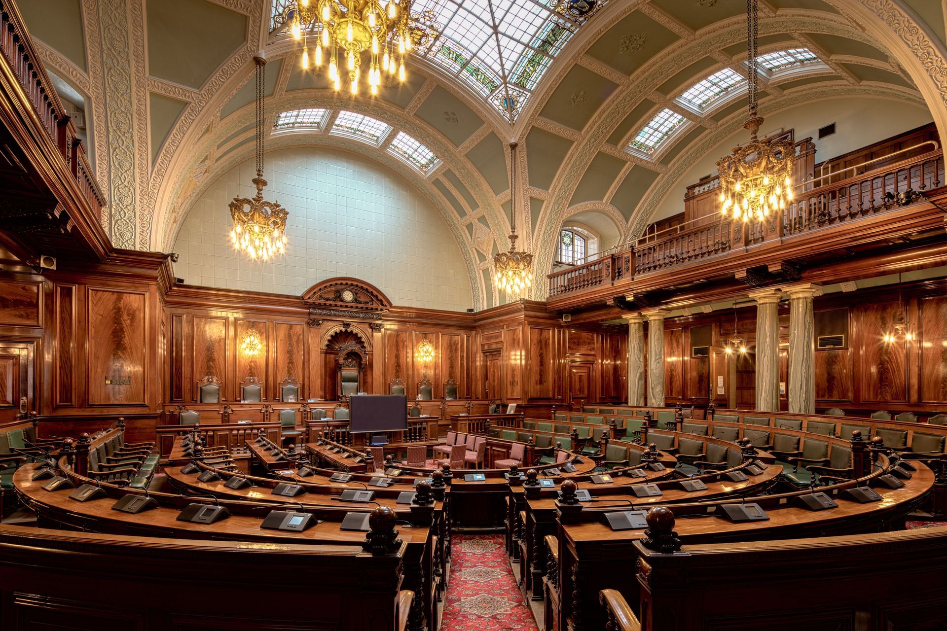 Grand ornate legislative chamber with carved wood paneling, arched ceiling, chandeliers, and rows of desks