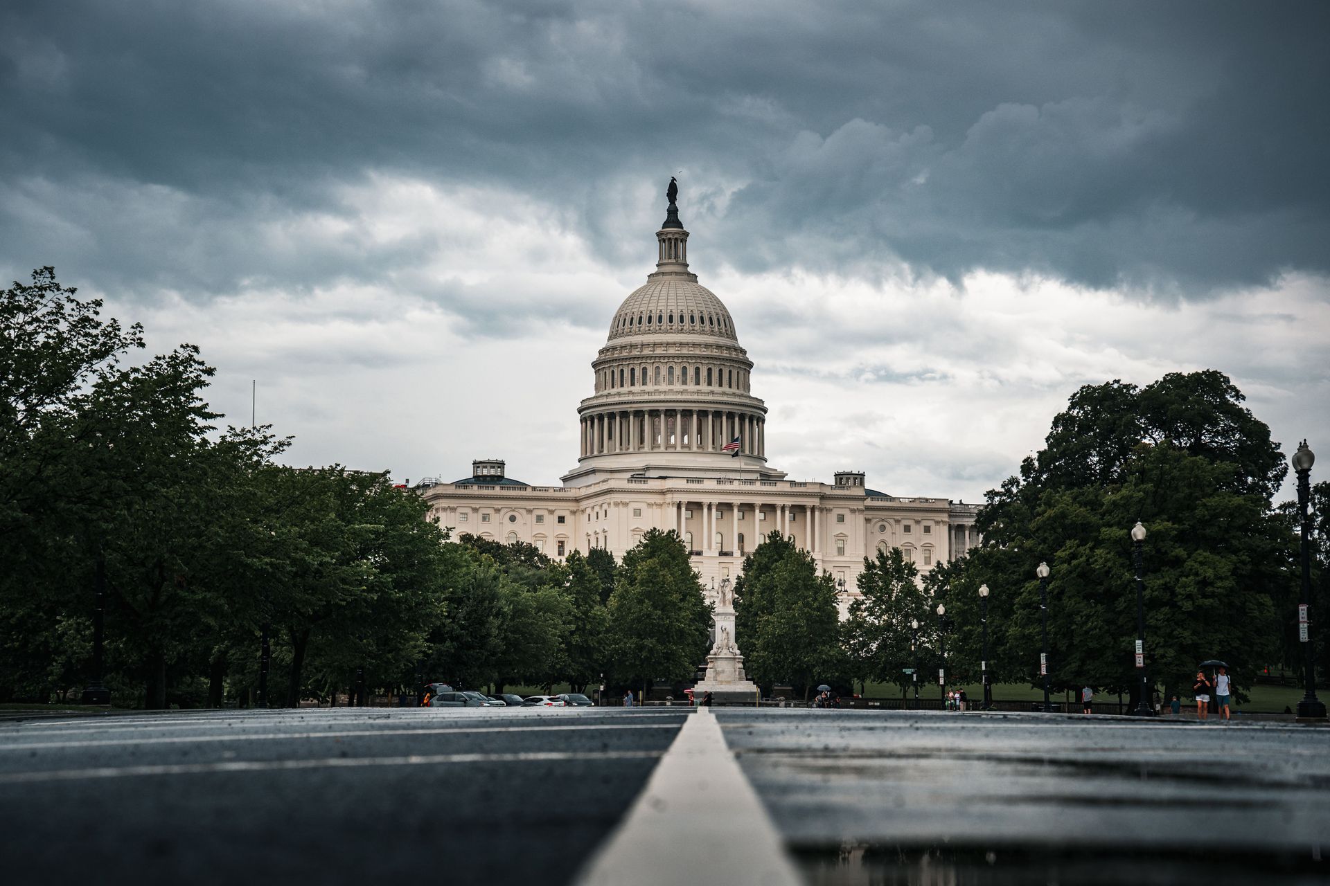 U.S. Capitol building framed by trees under a cloudy sky, seen from a wet road centerline