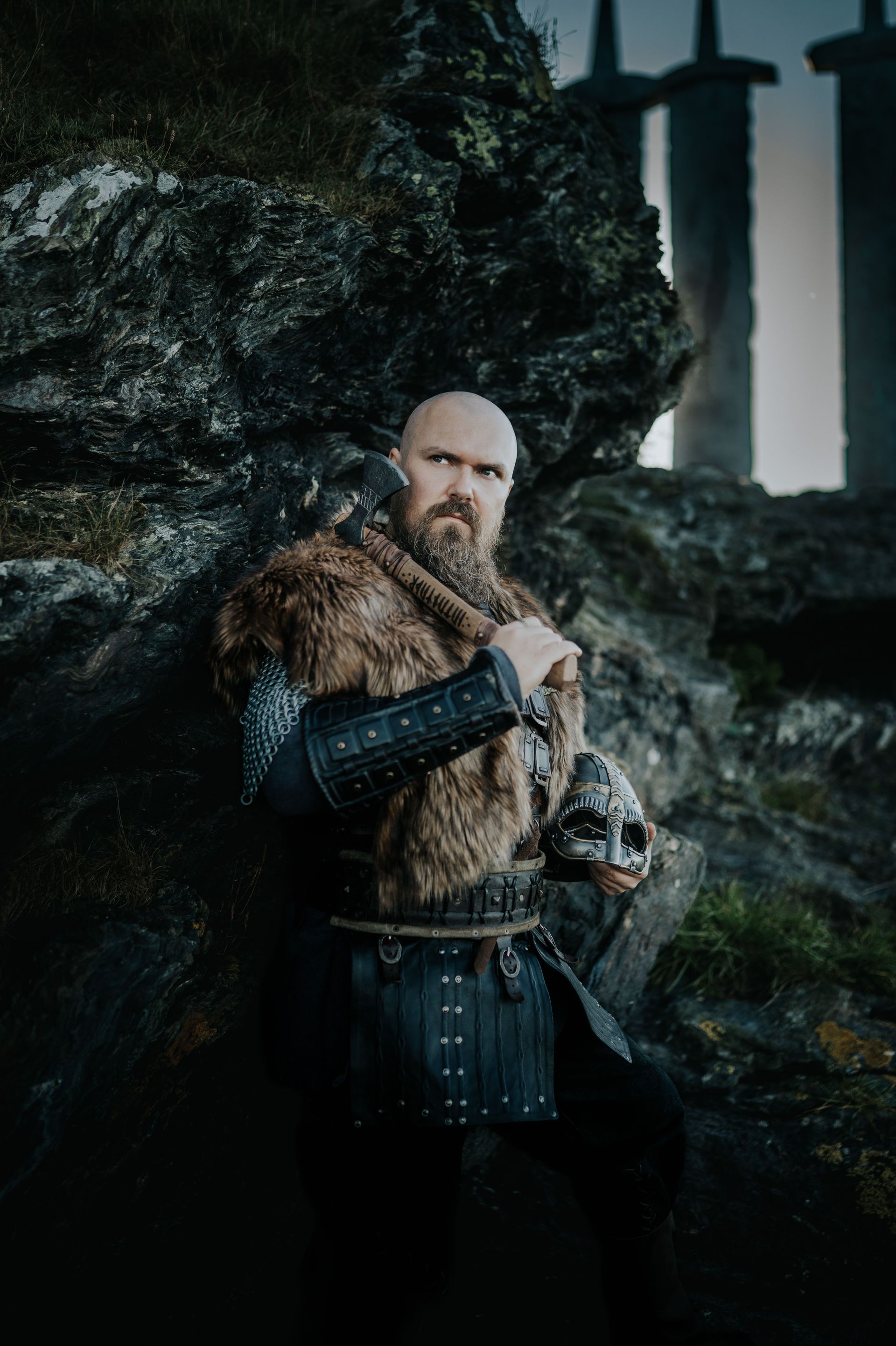 Bearded warrior in fur cloak holding a sword in a dark forest beside stone pillars