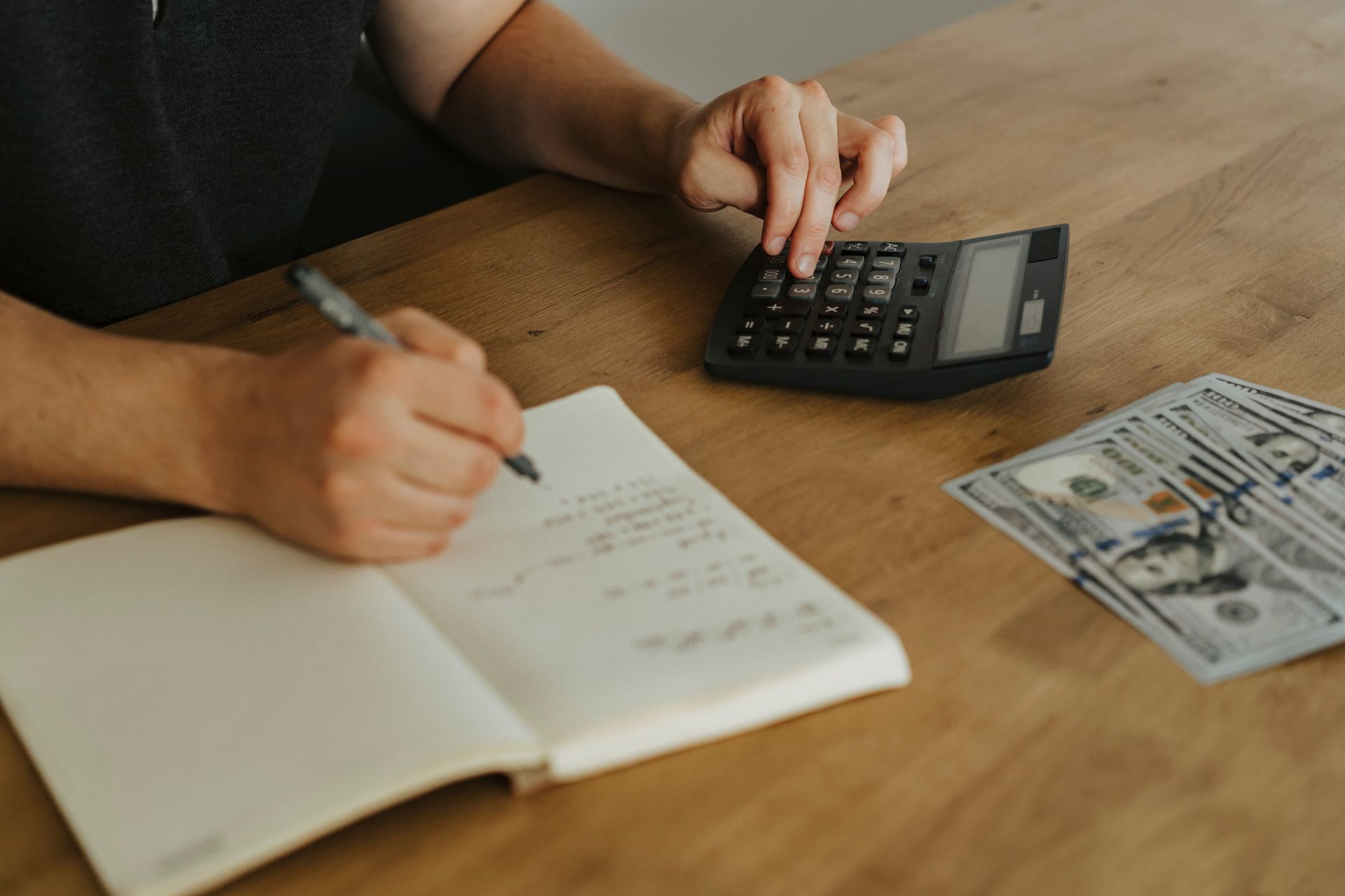 Hands writing in a notebook beside a calculator and paper money on a wooden table.
