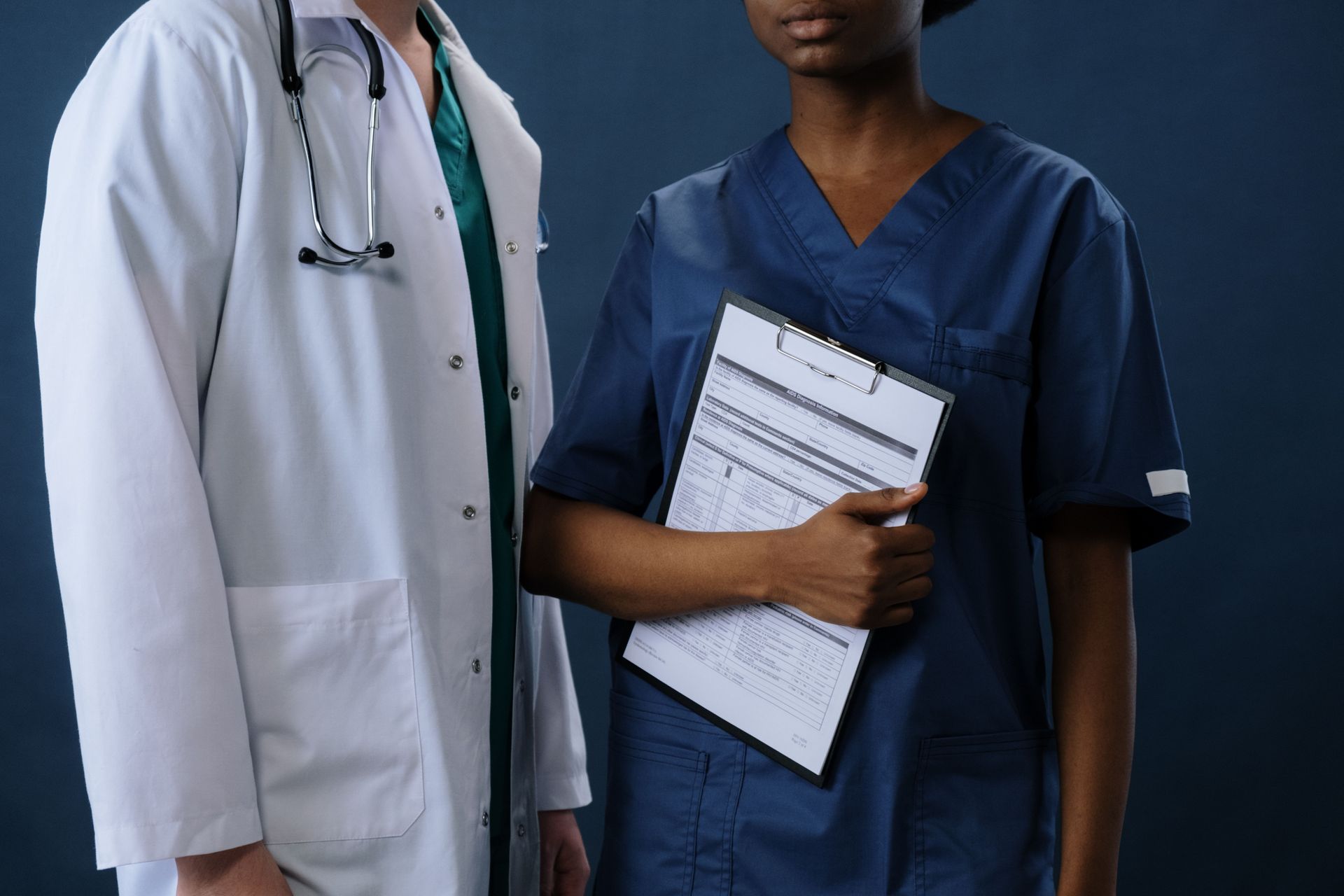 Two healthcare workers in scrubs and a white coat, one holding a clipboard