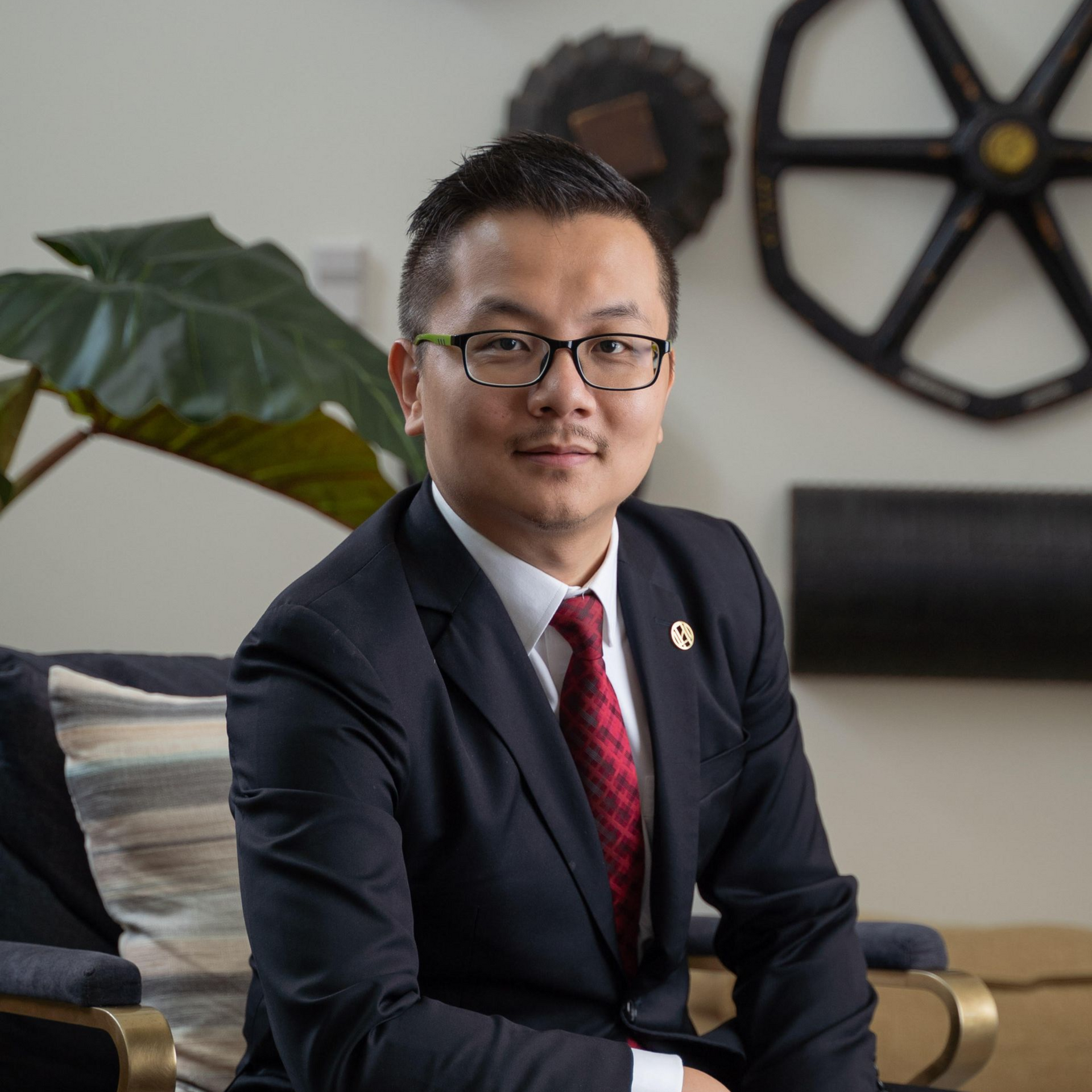 Man in suit and red tie sitting indoors with plant and wall decor in the background
