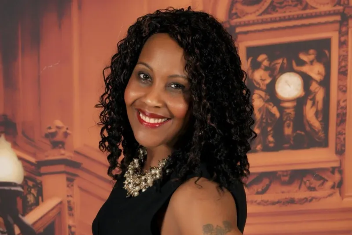 Smiling woman in a black dress with curly hair posing in front of a warm-toned decorative backdrop