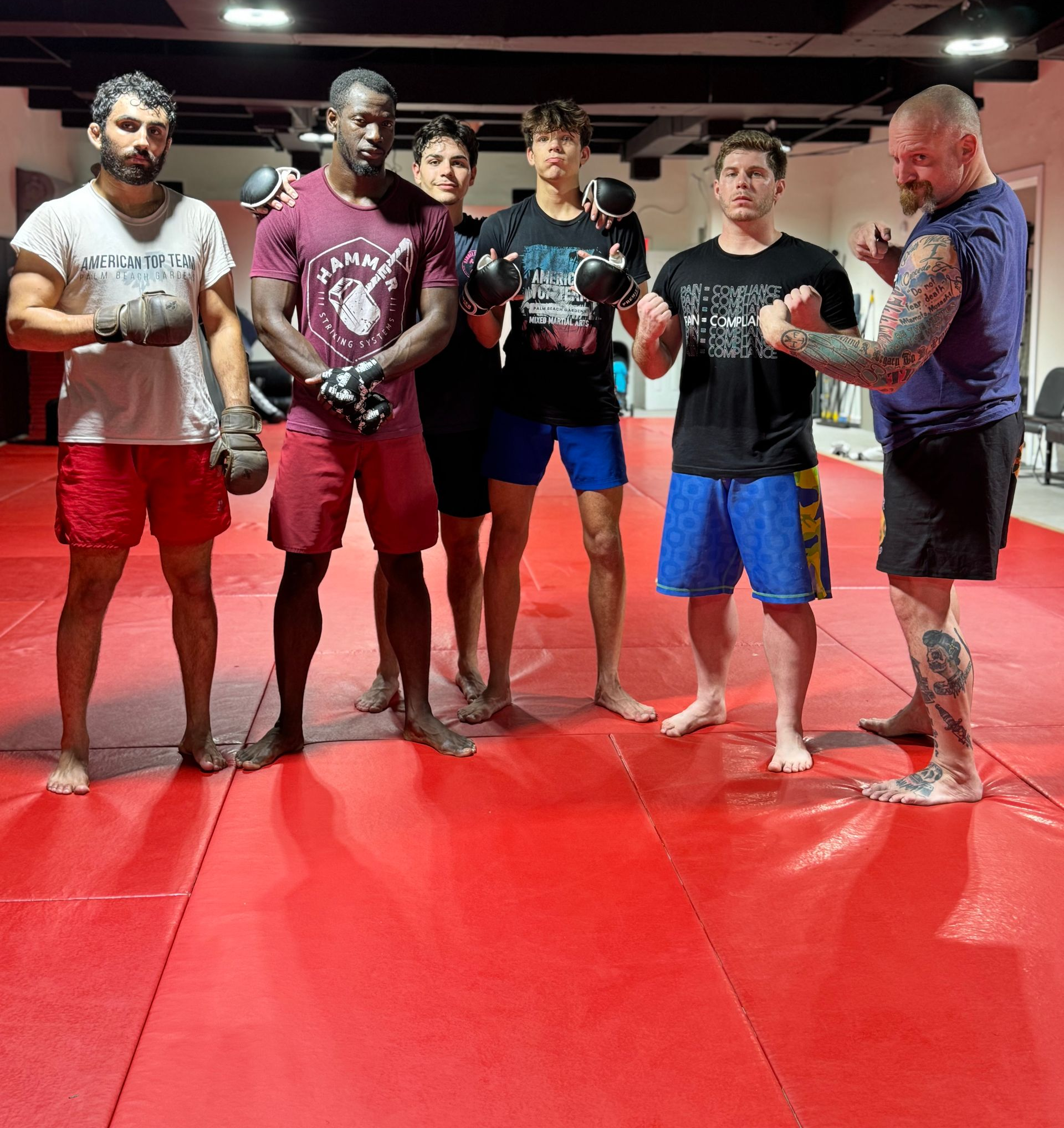 A group of people posing together in a fitness studio, most making hand gestures, with others training in the background.