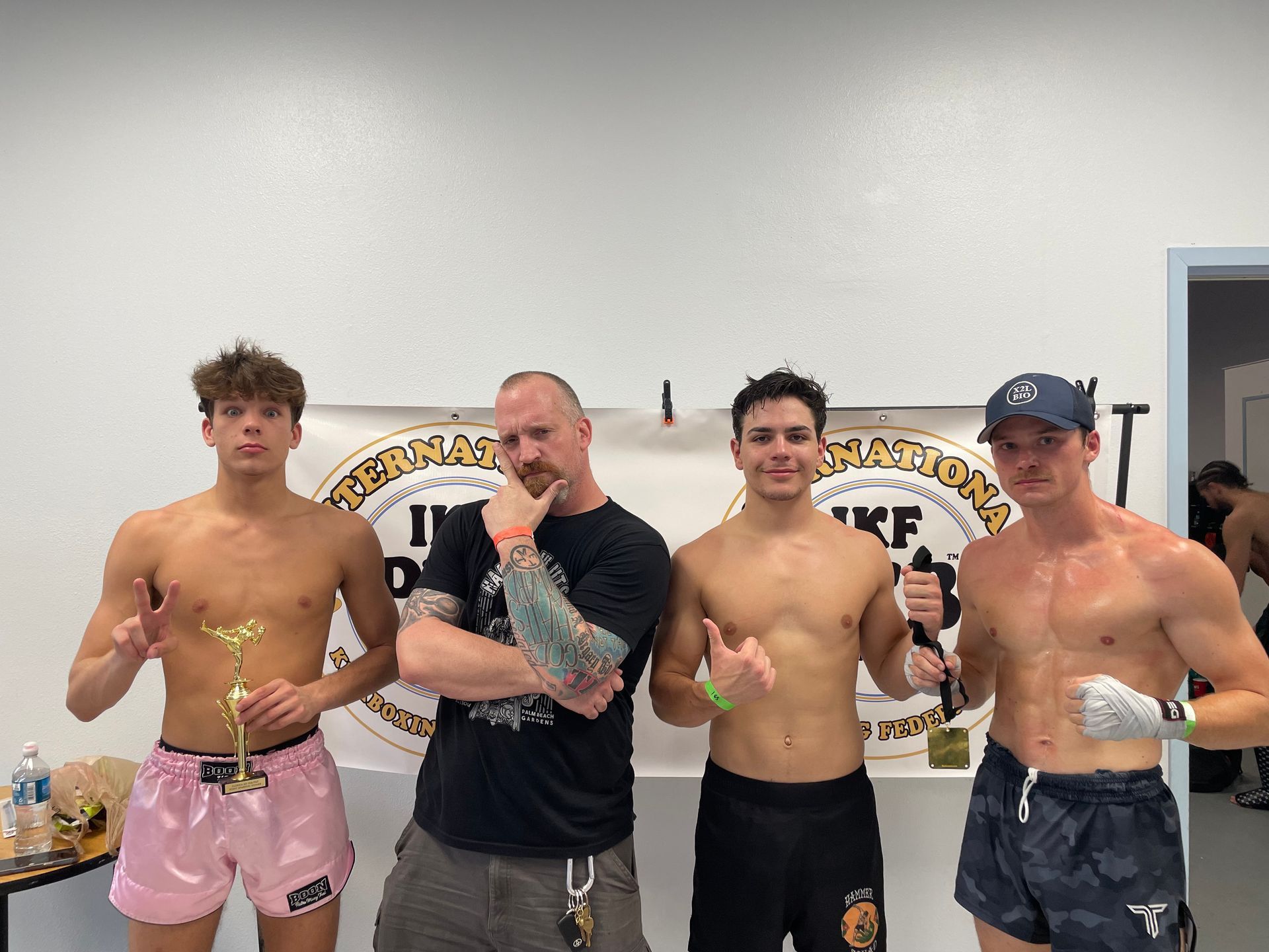 Four shirtless men holding trophies pose in a gym with a grappling club banner behind them.