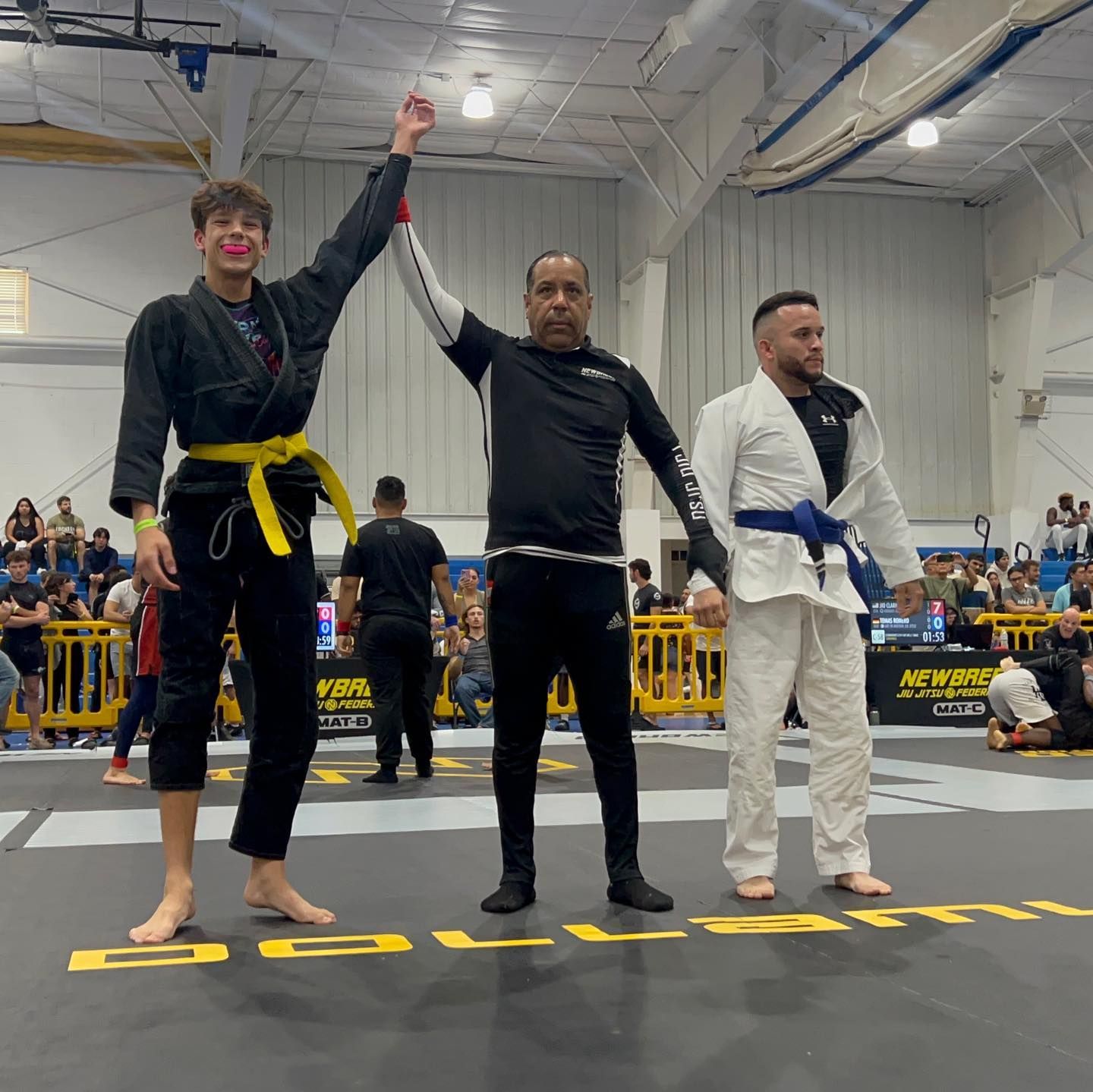 Three men are standing on a wrestling mat and one of them is wearing a yellow belt