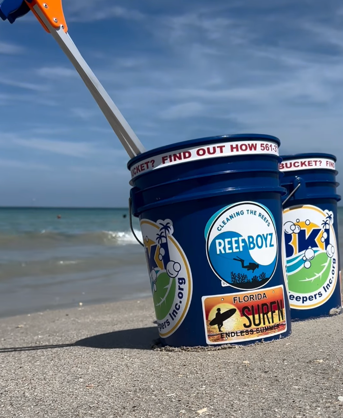Two blue buckets with stickers on a sandy beach, a grabbing tool overhead, ocean in background.