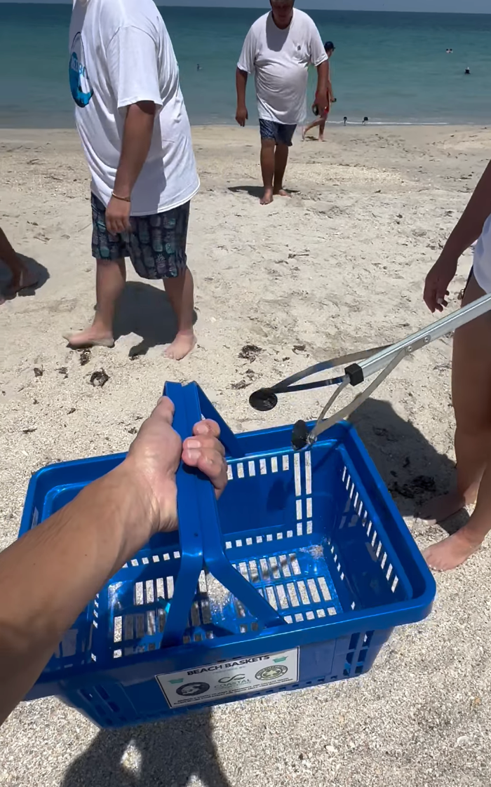 Person holding a blue basket and grabber picking up trash on a sandy beach with others walking in the background.