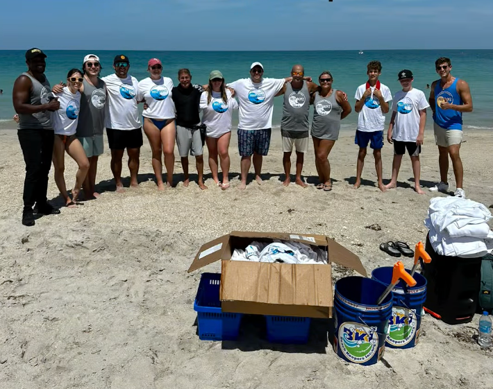 Group of volunteers on a beach, smiling and posing with cleanup supplies. Box of shirts, buckets, and the ocean in the background.