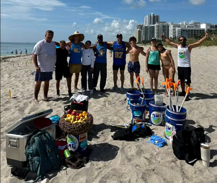 A group of people stand on a sandy beach, smiling, next to cleaning supplies and a basket of fruit, with buildings and ocean in the background.