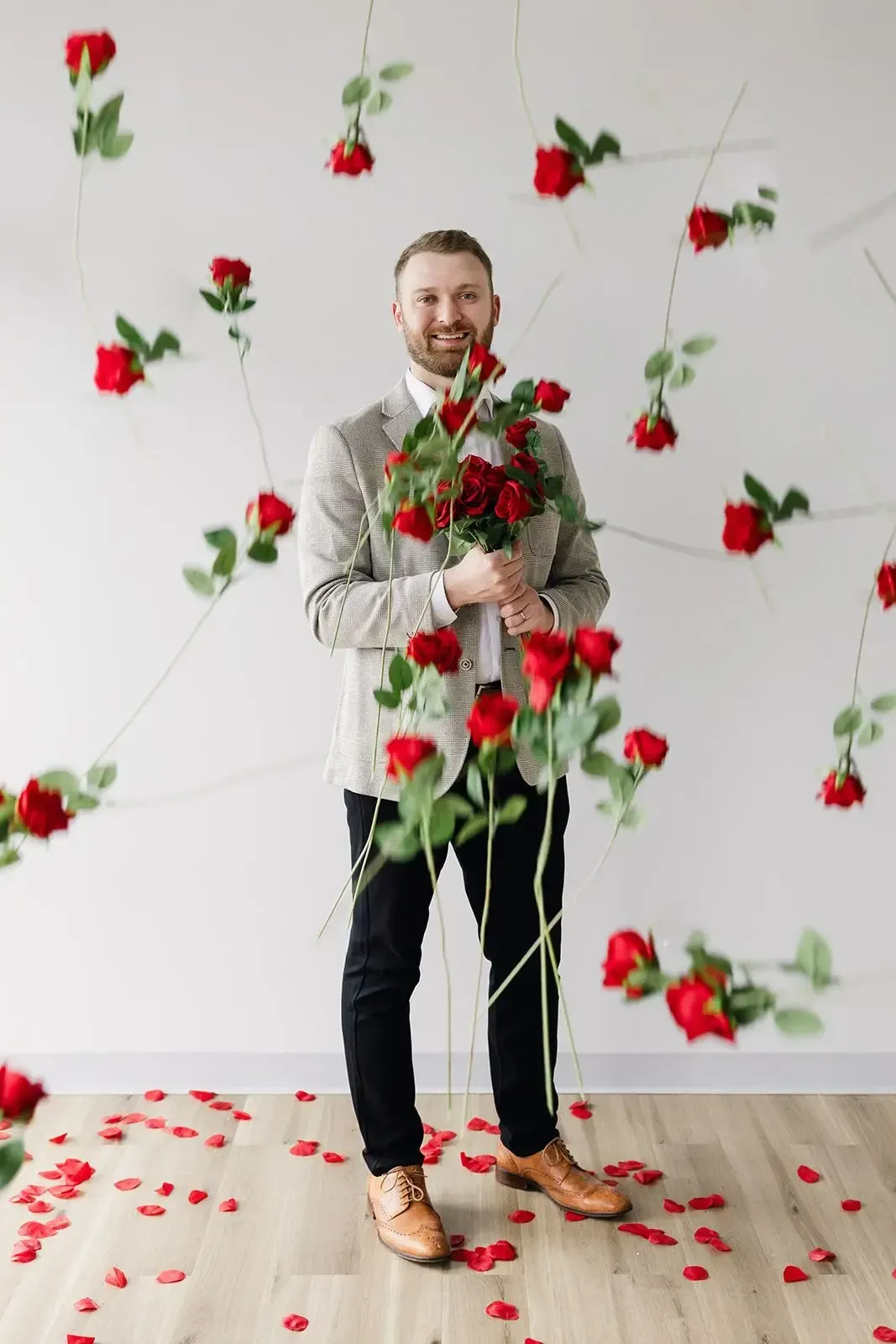 A man is holding a bouquet of red roses in front of a wall of red roses.
