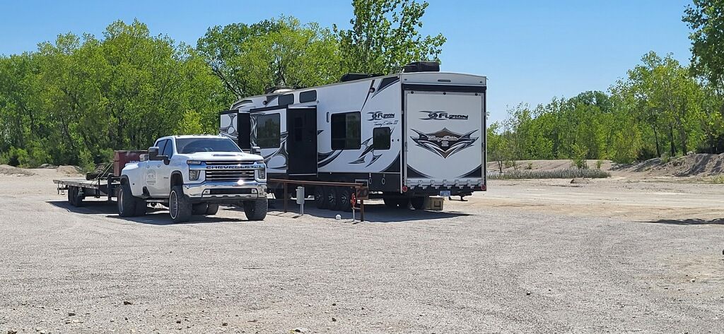 White pickup truck parked beside a black-and-white RV in a gravel lot near trees under a clear sky