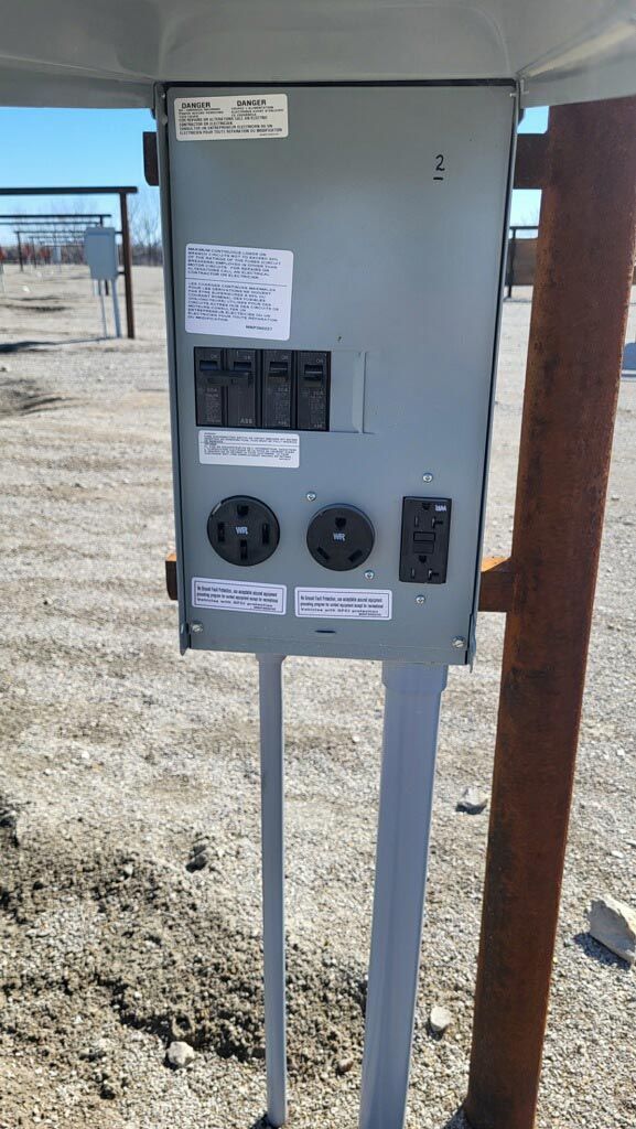 Gray electrical control panel mounted outdoors on metal posts under a canopy, with dials and labels.