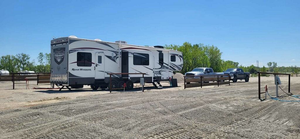 RVs and pickup trucks parked on a gravel lot under a clear blue sky, with trees in the background