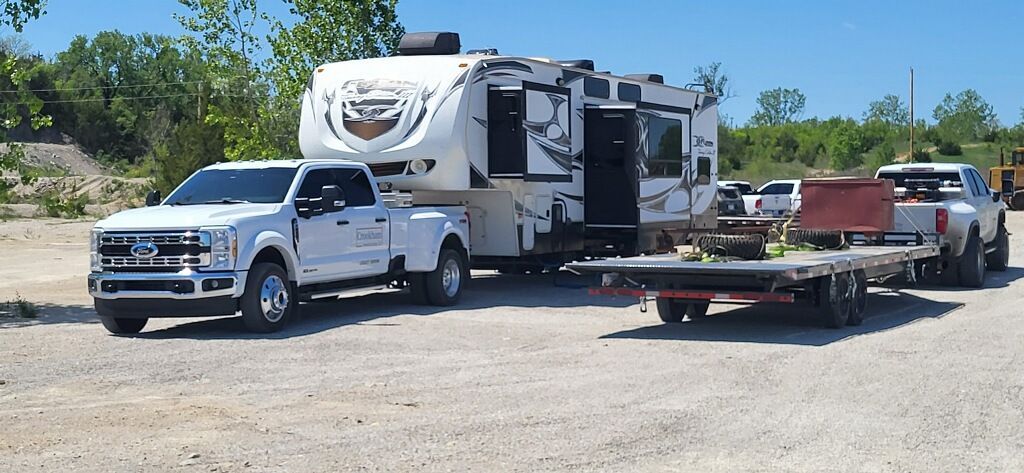 White truck towing a large camper trailer with a flatbed trailer behind, parked on a gravel lot under blue sky