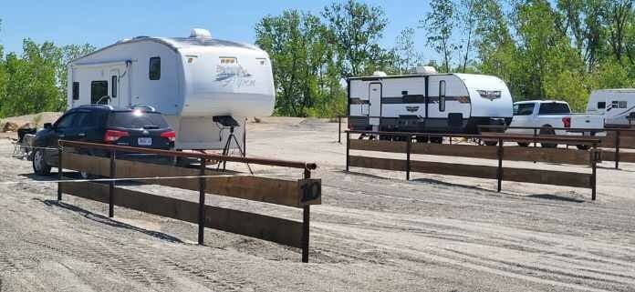 RVs and trailers parked in a dusty lot behind rust-colored fencing under a clear sky
