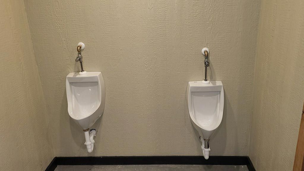 Two white wall-mounted urinals in a beige restroom with a dark floor trim at Workforce RV Park Rayville, MO