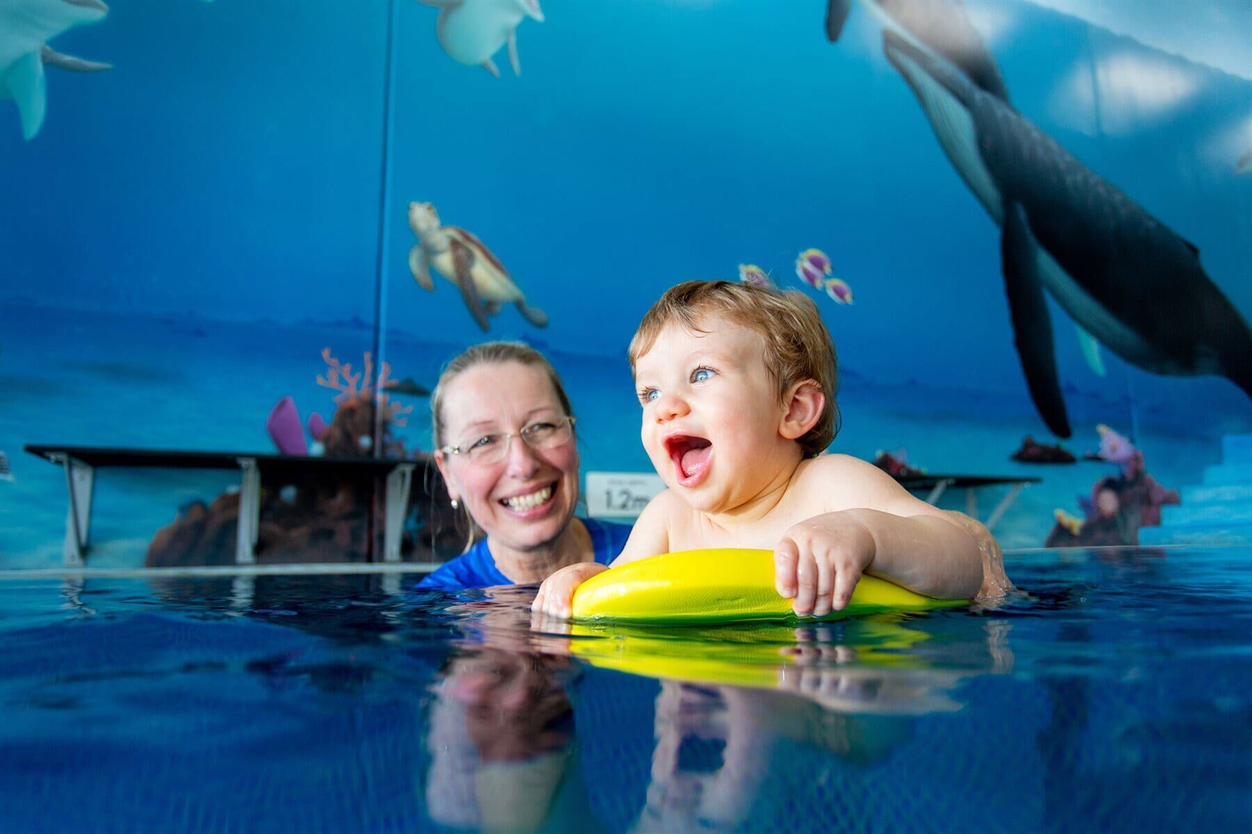 Woman Smiles, Holding a Yellow Float with A Baby in A Pool with Ocean-Themed Mural Background — Swim Care Boambee in Coffs Harbour, NSW