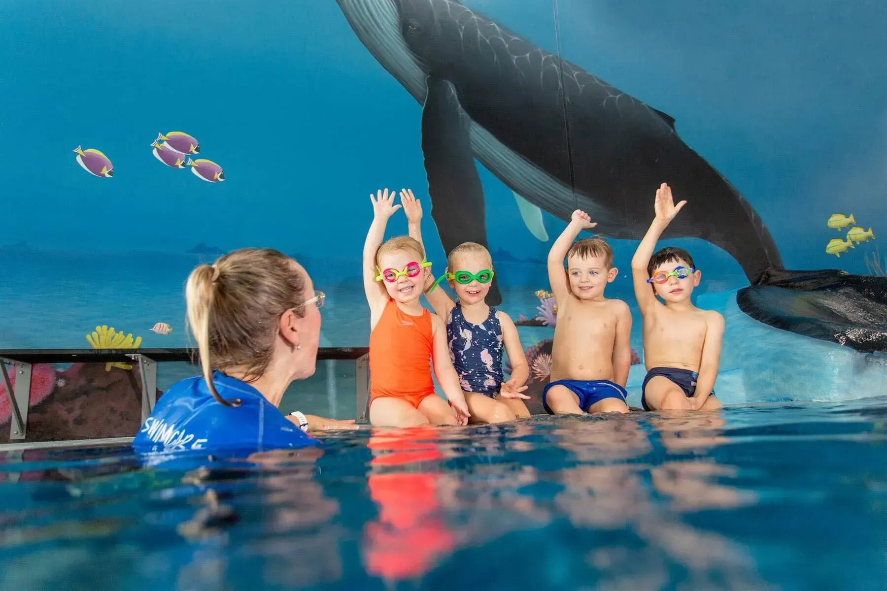 Four young children in goggles sitting poolside during a swim lesson, with a swim instructor.