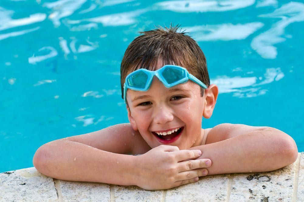 Boy in Swimming Goggles Smiles in A Pool, Resting Arms on The Edge of The Pool — Swim Care Boambee in Coffs Harbour, NSW