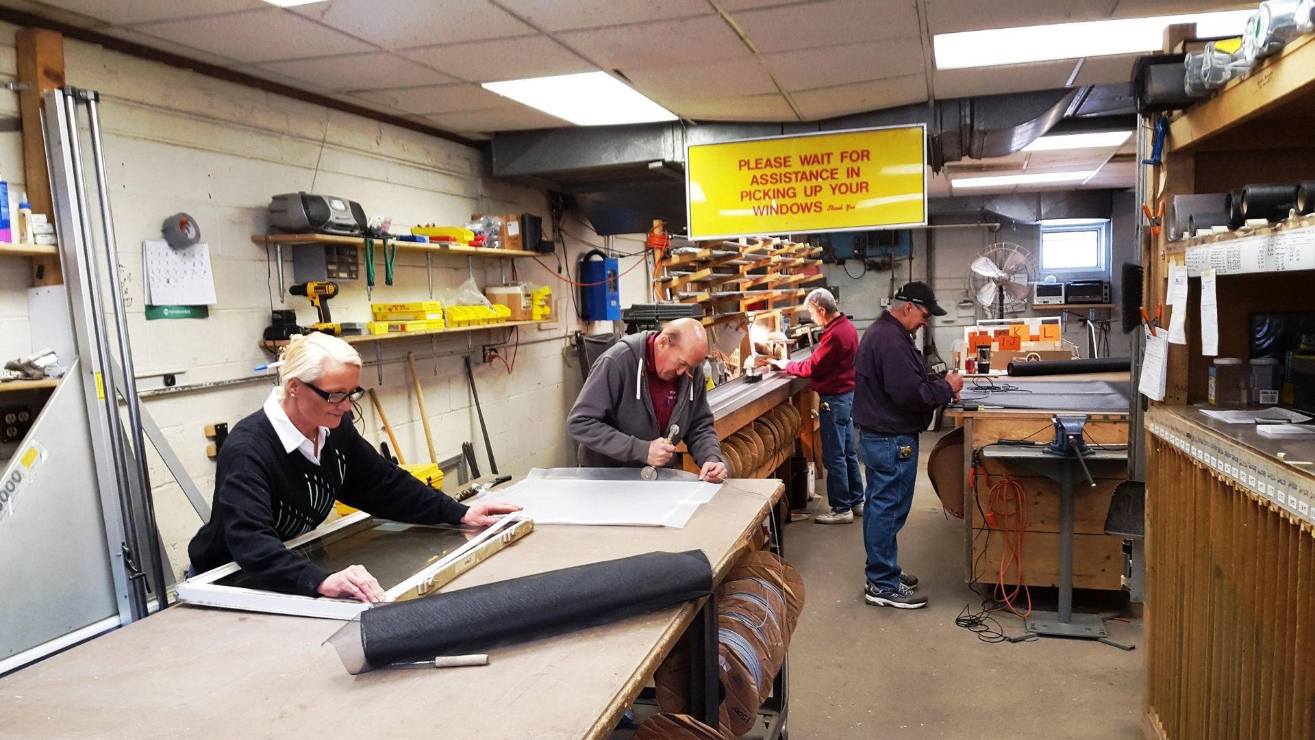 Four people working at workbenches in a workshop with tools and supplies. Four people working at workbenches in a workshop with tools and supplies.