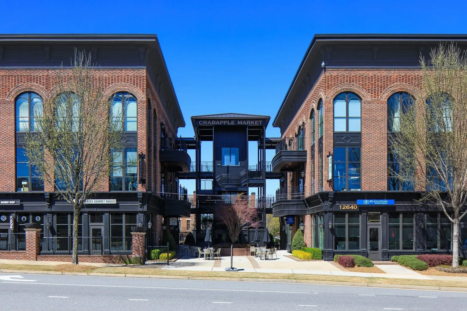 Two brick buildings flank a central courtyard, with large arched windows and a covered walkway under a blue sky.