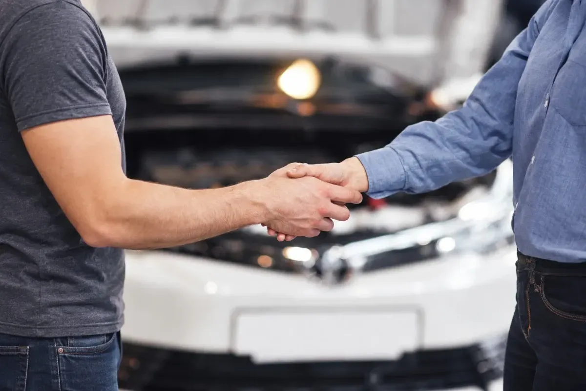 Two people shaking hands in front of a white car with its hood open