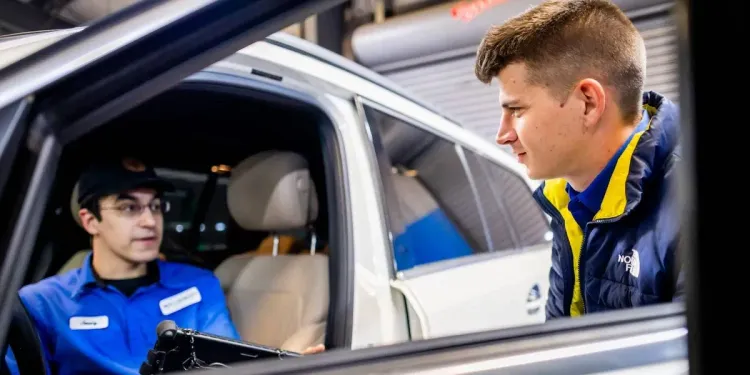 Two people in blue uniforms talk beside a white car, with one leaning in from outside the open window.