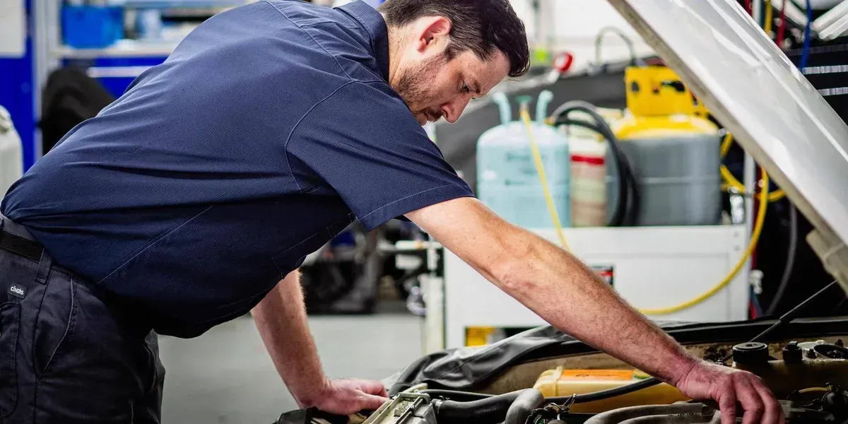 Mechanic in blue shirt working under a car in a garage, reaching into the engine bay.