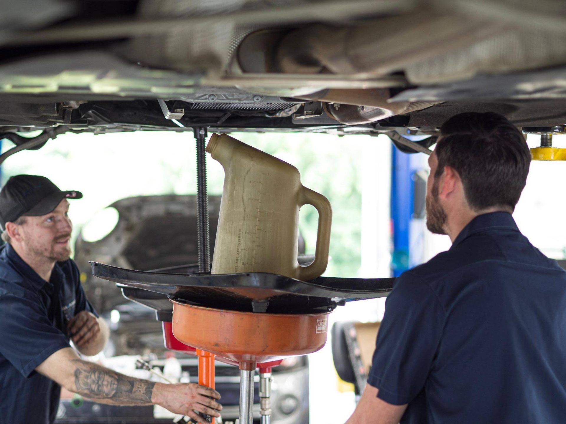 Two mechanics inspect a car on a lift, with an orange oil drain pan underneath.