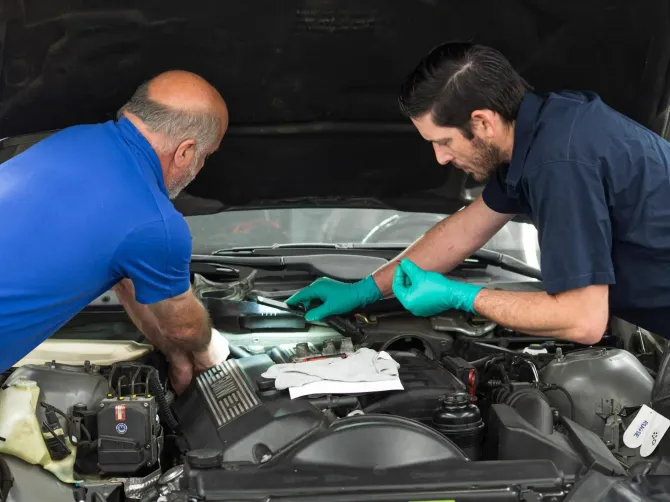 Two mechanics in blue and black shirts work under a car hood in a garage, wearing gloves.