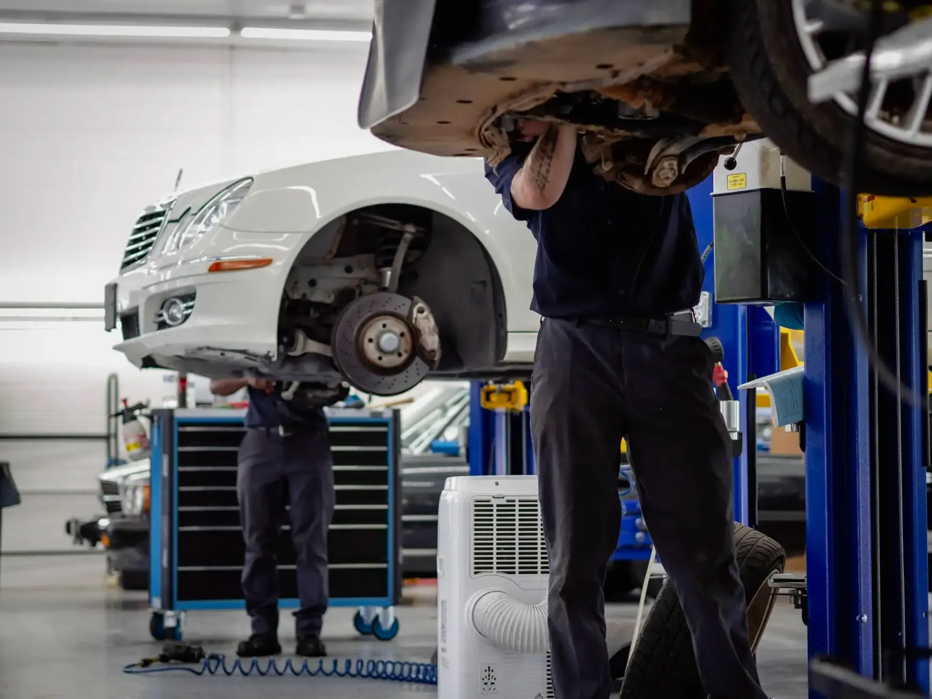 Mechanic working under a raised white car in an auto repair shop, with tools and equipment nearby