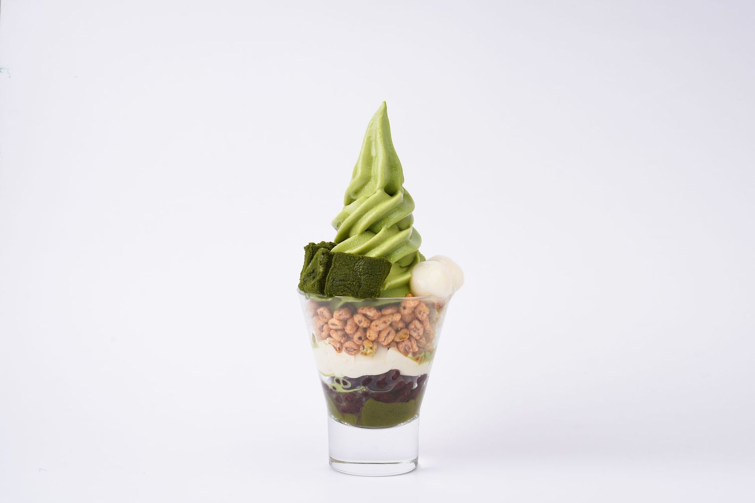 Tall dessert parfait with green soft-serve, nuts, whipped cream, and chocolate layers in a clear glass