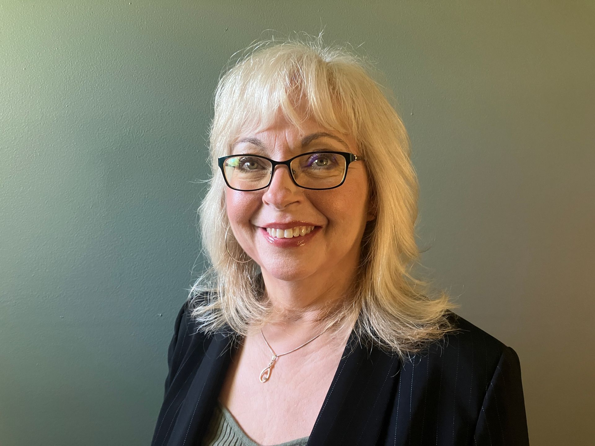 Smiling person with glasses and blonde hair in a black jacket against a gray background