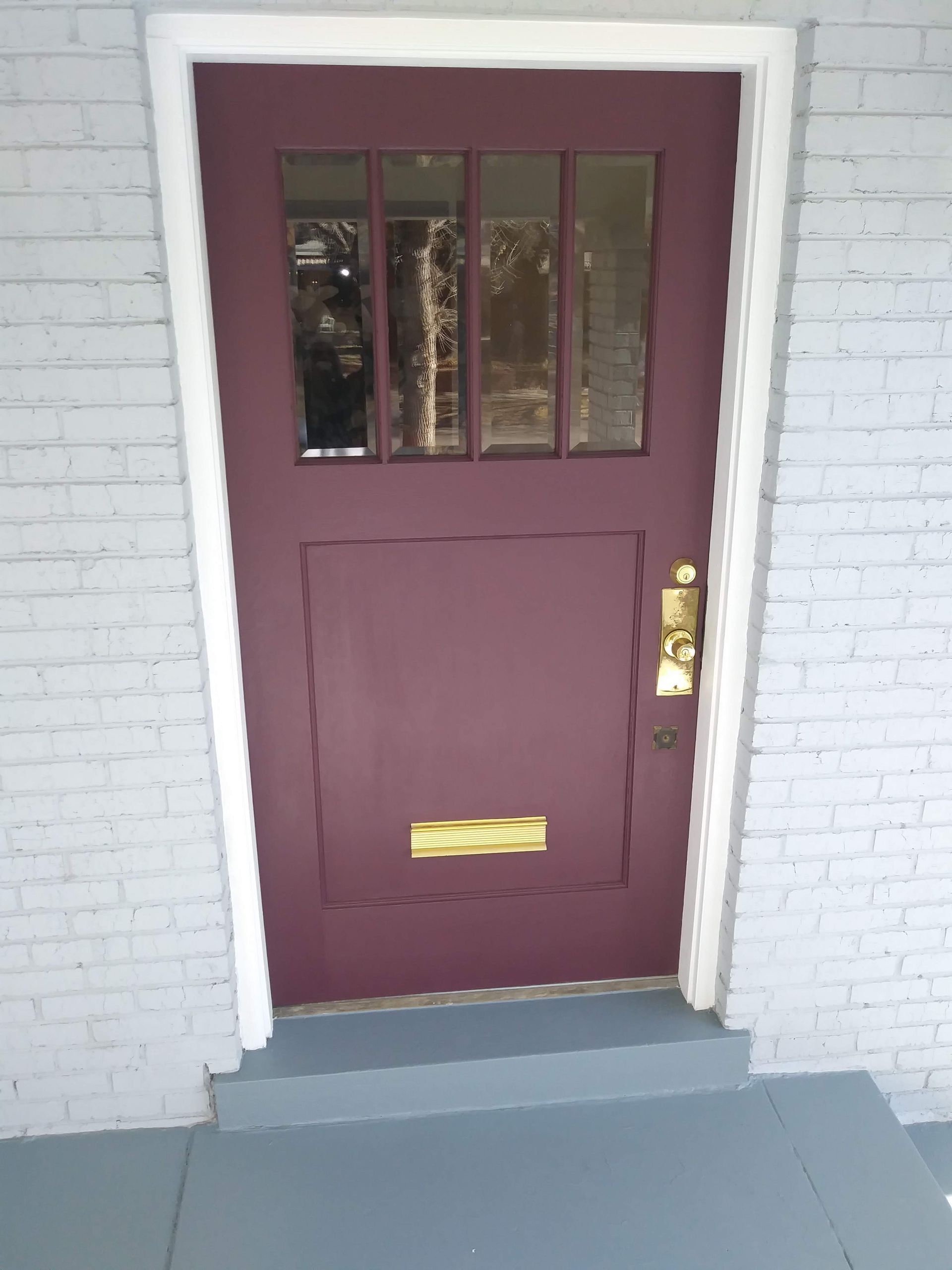 Maroon front door with brass mail slot and glass panes in a white brick entryway