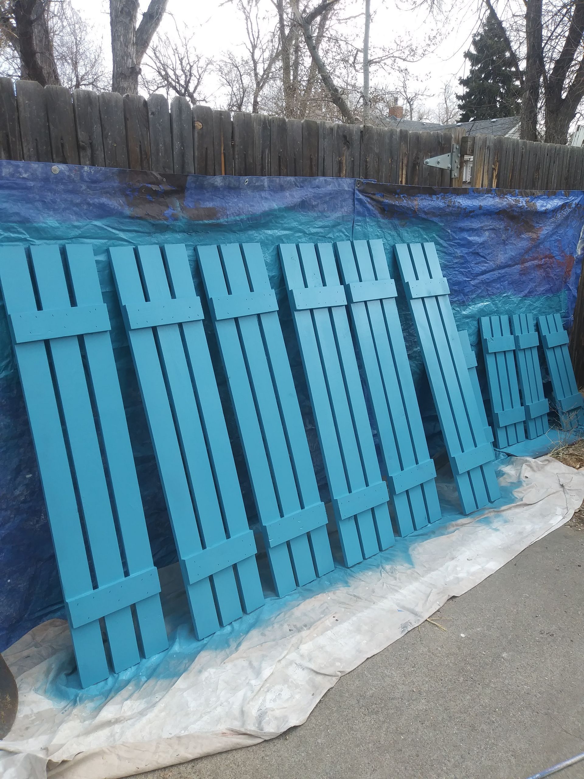 Stacked turquoise wooden pallets leaning against a blue tarp outdoors on pavement.
