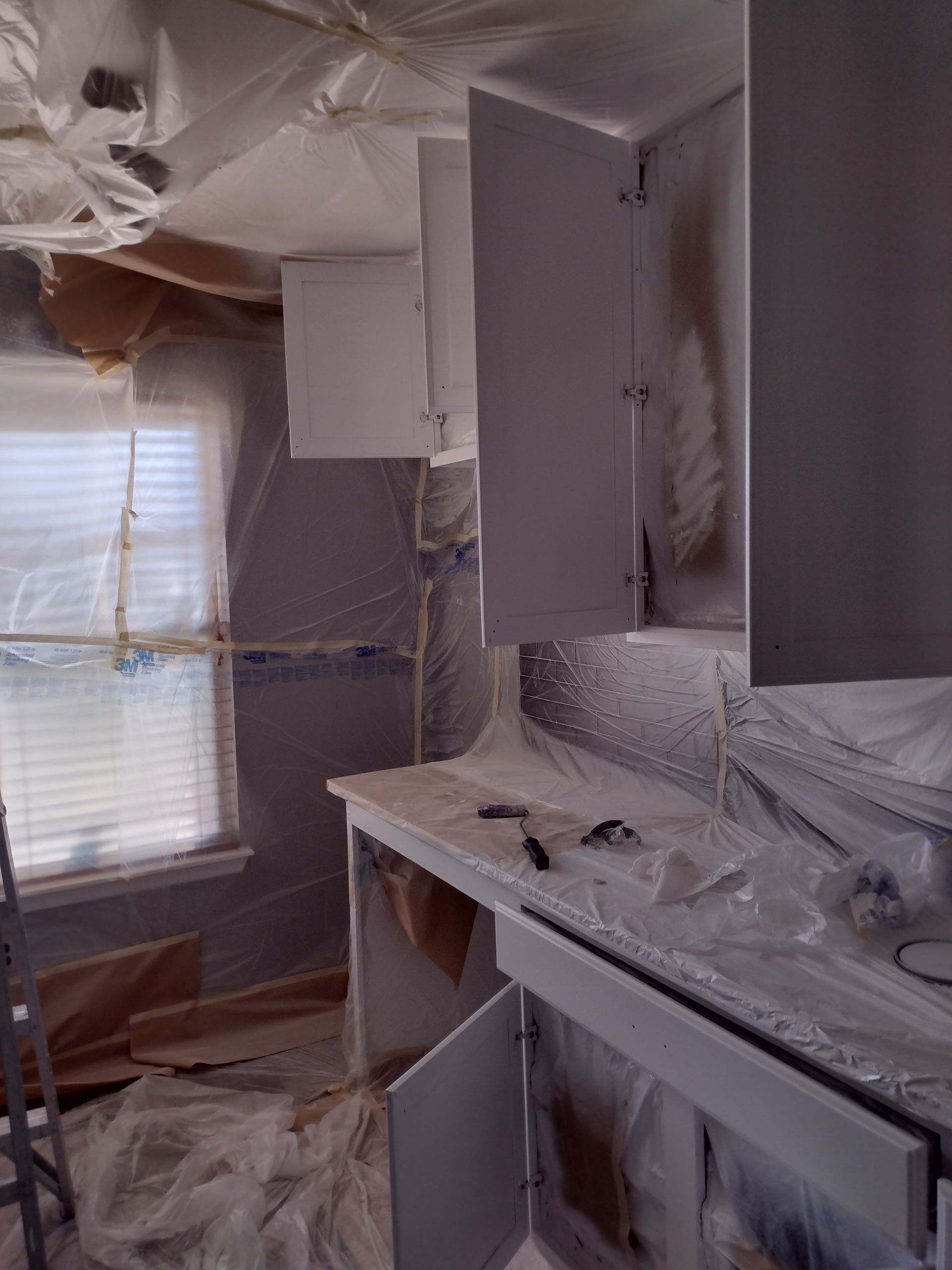 Kitchen covered in plastic sheeting and dust, with cabinets, countertop, and window during renovation