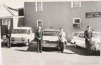 Black-and-white photo of three men standing beside classic cars outside a building