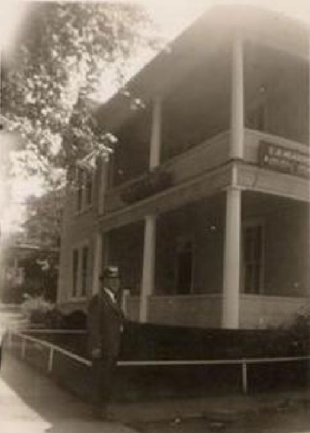 Old two-story house with a wraparound porch, seen from the front with a person standing by the steps.