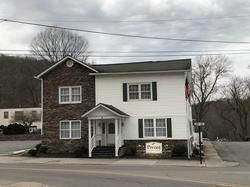 Small two-story white and stone building with a porch and sign along a road on an overcast day