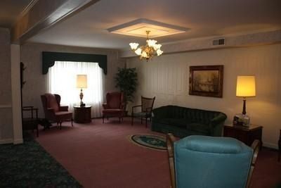 Dim living room with green sofa, blue chair, lamps, chairs, and a ceiling light on maroon carpet.