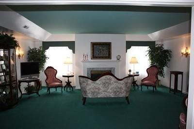 Living room with green carpet, fireplace, ornate chairs, and potted plants in a symmetrical arrangement