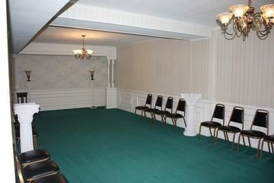 Empty banquet hall with green carpet, white paneled walls, chandeliers, and rows of black chairs along the side