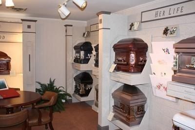 Display of ornate wooden coffins in a showroom, with shelves, plants, and a “Her Life” sign on the wall