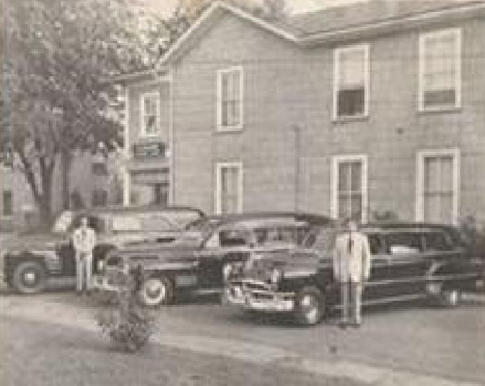 Vintage black-and-white photo of three classic cars parked beside a building with two people standing nearby.