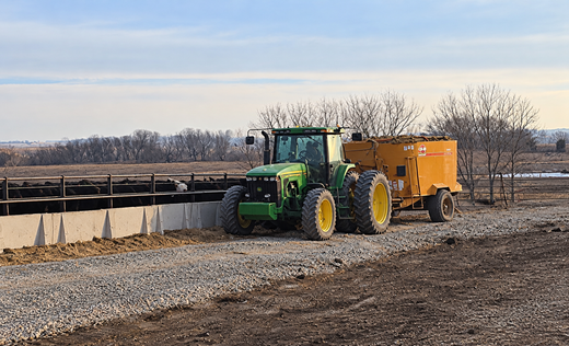 Green tractor towing a yellow manure spreader beside a fenced field on a rural farm road