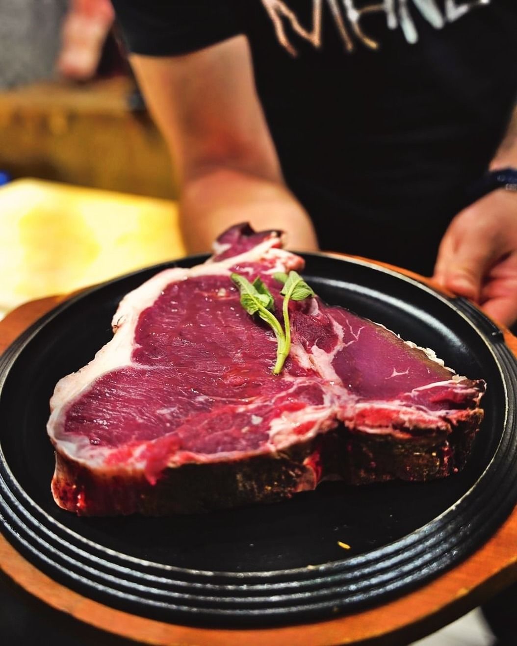 Una grande bistecca alla fiorentina cruda su un piatto nero, tenuta da una persona.