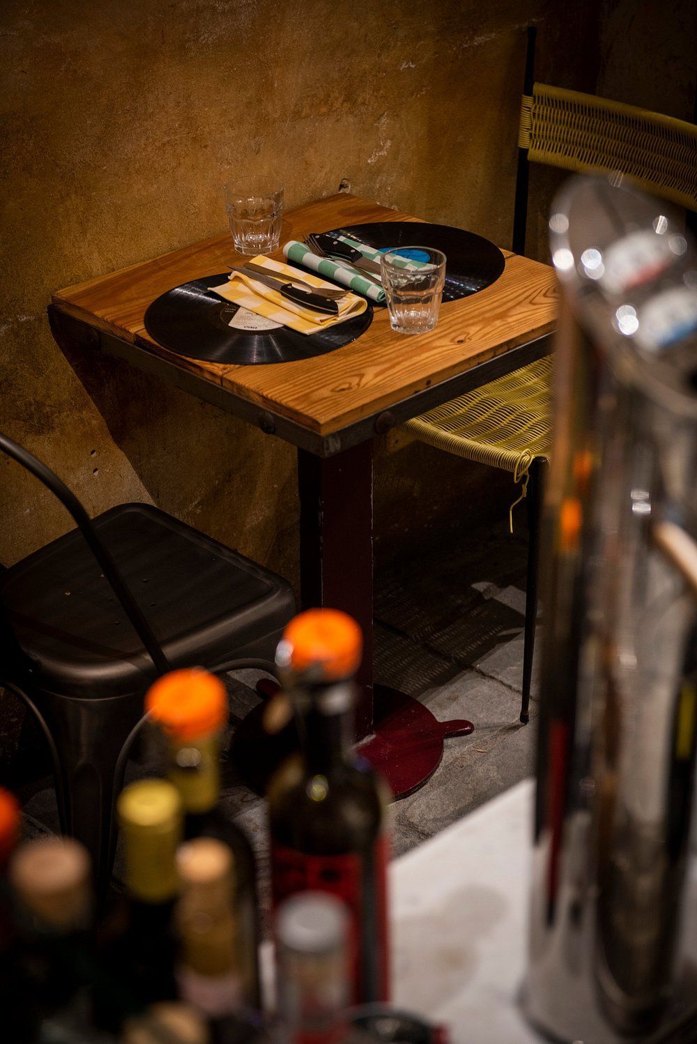 Un tavolino di legno con ripiani in vinile, due bicchieri, bottiglie e una sedia di metallo, in un bar scarsamente illuminato.